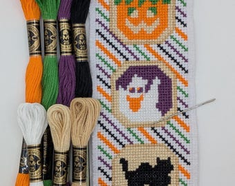 May include: A Halloween-themed cross-stitch project featuring a pumpkin, ghost, and black cat design. The project includes a needle and skeins of embroidery floss in orange, green, purple, black, white, and tan colors. The background has diagonal stripes.