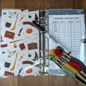 May include: A binder with a clear cover, featuring a whimsical pattern of magical elements like owls, potions, and wands. Inside, a thread inventory sheet and a wet erase marker are visible, along with colorful embroidery floss.