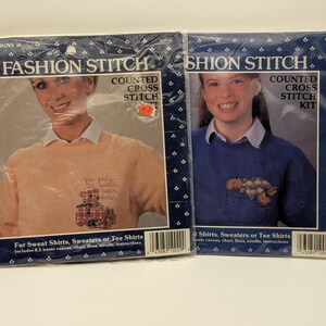 May include: Two "Fashion Stitch" counted cross stitch kits. One kit features a woman wearing a peach-colored sweater with a teddy bear design. The other kit shows a woman in a blue sweater with a floral design.