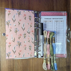 May include: An open clear binder with a floral patterned cover and a thread inventory sheet. Inside are several skeins of embroidery floss in various colors, a black marker, and a white marker.