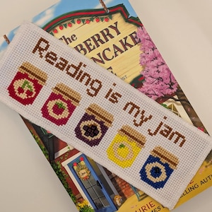 May include: A white cross-stitch bookmark with the text "Reading is my jam" and images of jam jars. The bookmark rests on a book titled "The Berry Pancake" by Laurie, a New York Times bestseller.