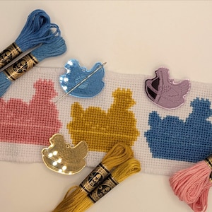 May include: A cross-stitch project featuring a rooster design in pink, yellow, and blue, with matching embroidery floss and rooster-shaped needle minders. The floss is in shades of blue, pink, and yellow. A needle is placed on the blue needle minder.