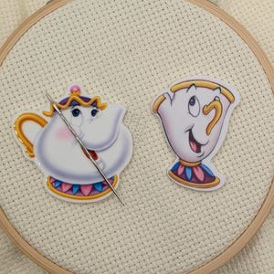 May include: Two cartoon character patches, a teapot and a teacup, are displayed on a white fabric stretched within a wooden embroidery hoop. A needle is inserted through the teapot patch. The teapot is white with a pink and blue design. The teacup is white with a gold rim.