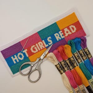 May include: A rectangular cross-stitch project on white Aida cloth featuring the text HOT GIRLS READ in white letters against a background of purple, blue, orange, and pink blocks. A silver needle, silver scissors, and eight embroidery floss skeins.