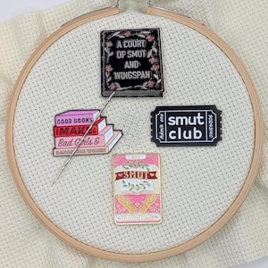 May include: Four enamel pins with book and ticket designs are displayed on a white fabric stretched in a wooden embroidery hoop. The pins feature text like "A Court of Smut and Wingspan" and "Smut Club."