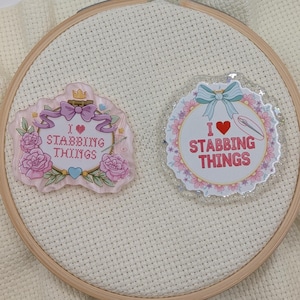 May include: Two decorative pins with the text "I ❤️ Stabbing Things". One pin is pink with roses and a crown, the other is a circle with a floral border and a needle. Both pins are on a white fabric background.