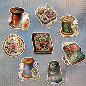 May include: A collection of eight stickers featuring sewing supplies: a red thread spool, a floral tin with scissors, a green thread spool, a floral embroidery hoop, a book titled Embroidery Lessons, a sewing kit with needles, a blue thread spool, and a silver thimble.