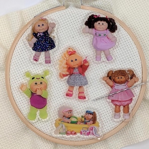 May include: A cross-stitch hoop featuring six Cabbage Patch Kids-themed acrylic charms. The charms depict dolls in various outfits, including dresses and a frog costume. The hoop is set against a white, textured fabric background.
