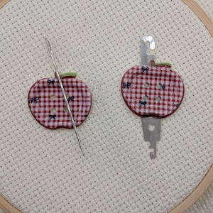 May include: Two apple-shaped needle minders and a needle on a white fabric background. The minders are red and white checkered with green stems and small bow accents. One minder has a metal needle holder attached.