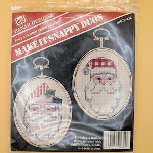 May include: A sealed package containing a cross-stitch kit. The kit features two oval frames with cross-stitched Santa Claus designs. The package reads "MAKE IT SNAPPY DUOS" and includes frames, floss, Aida fabric, charts, needle, and instructions.