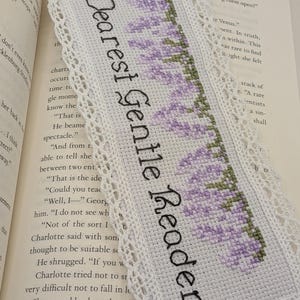 May include: White cross-stitch bookmark with the text "Dearest Gentle Reader" in black. The bookmark features a lavender floral design and a delicate lace trim. The bookmark is resting on the pages of a book.