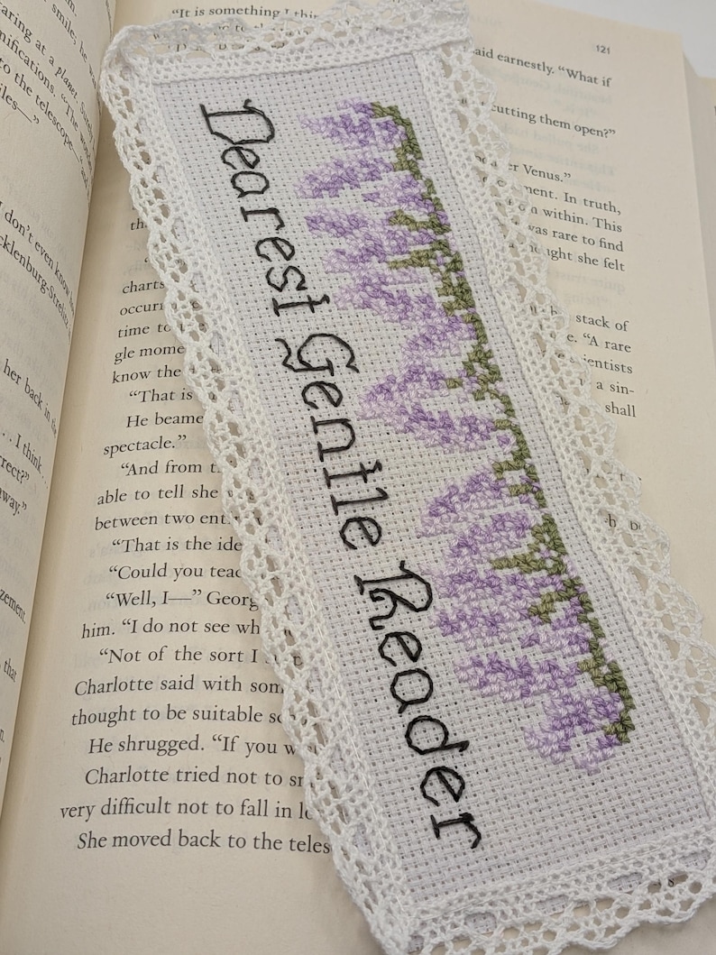 K&ouml;nnte beinhalten: Wei&szlig;es Kreuzstich-Lesezeichen mit dem Text "Dearest Gentle Reader" in Schwarz. Das Lesezeichen hat ein Lavendel-Blumenmuster und eine zarte Spitzenborte. Das Lesezeichen liegt auf den Seiten eines Buches.