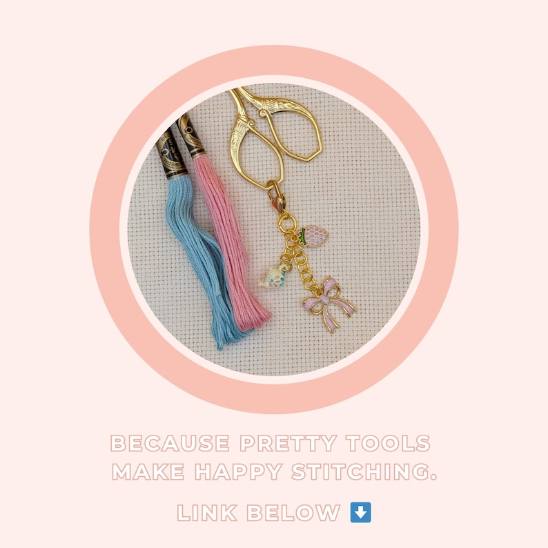 Peut inclure: Gros plan sur des outils et fournitures de broderie. Des ciseaux dor&eacute;s sont attach&eacute;s &agrave; un charm avec un n&oelig;ud rose, une fraise et d'autres breloques. Deux &eacute;chevettes de fil &agrave; broder, une bleue et une rose, sont &eacute;galement visibles. Texte : BECAUSE PRETTY TOOLS MAKE HAPPY STITCHING.