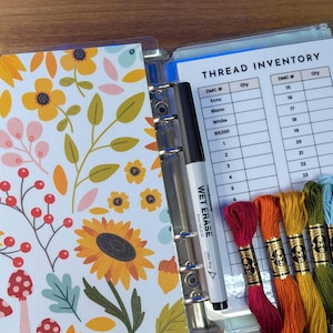 May include: A clear binder with a floral cover featuring sunflowers, leaves, and berries. Inside, a thread inventory sheet is visible, along with a black marker and several skeins of colorful embroidery floss. The text "THREAD INVENTORY" is visible.