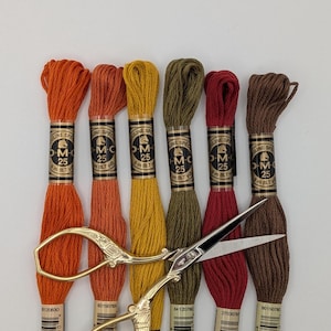 May include: Six skeins of DMC embroidery floss in autumn-inspired shades of orange, yellow, olive green, red, and brown, each with a paper label marked 8m (8.7 yd). A pair of ornate gold-toned metal scissors rests across the thread skeins.