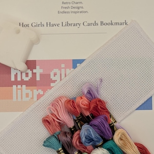 May include: A cross-stitch kit featuring a white Aida cloth strip, a white plastic thread bobbin, and a collection of multi-colored embroidery floss skeins. The pattern instruction sheet displays the text Hot Girls Have Library Cards Bookmark.