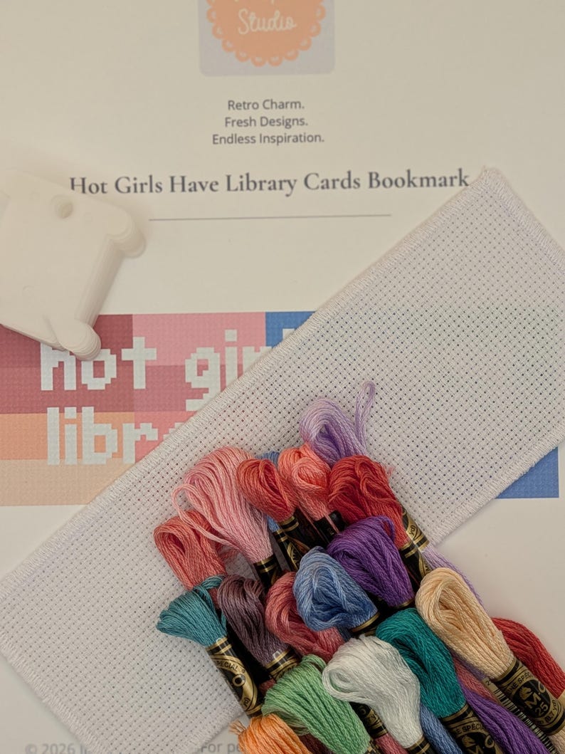 May include: A cross-stitch kit featuring a white Aida cloth strip, a white plastic thread bobbin, and a collection of multi-colored embroidery floss skeins. The pattern instruction sheet displays the text Hot Girls Have Library Cards Bookmark.