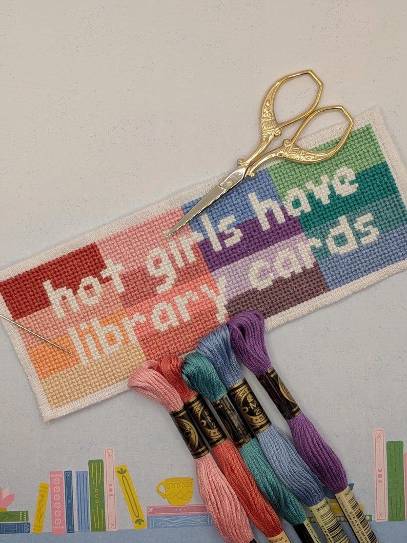 May include: A rectangular cross-stitch project featuring the text "hot girls have library cards" in white against a background of color-blocked squares in red, pink, purple, and teal. Gold-toned scissors and a needle rest on the fabric.