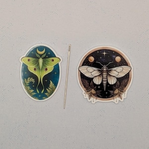 May include: Two circular stickers featuring moth illustrations and a thin metal needle. The left sticker shows a green luna moth on a dark blue background. The right sticker displays a white moth on a black background with celestial elements.