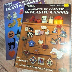 May include: Two Leisure Arts craft instruction booklets titled Holiday Magnets for Plastic Canvas and Magnets Go Country in Plastic Canvas. The covers feature colorful plastic canvas needlepoint designs including animals, holiday icons, and geometric patterns.