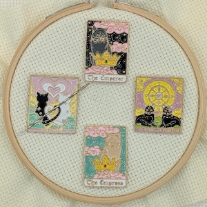 May include: Four enamel pins featuring tarot card designs with cat illustrations. The pins include "The Emperor," "The Lovers," "The Wheel of Fortune," and "The Empress." Each pin has a unique color scheme and design, set on a cream-colored fabric within an embroidery hoop.
