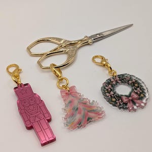 May include: A collection of Christmas-themed charms and a pair of gold scissors. The charms include a pink nutcracker, a Christmas tree with pink and white accents, and a wreath with pink bows. All charms have gold clasps.