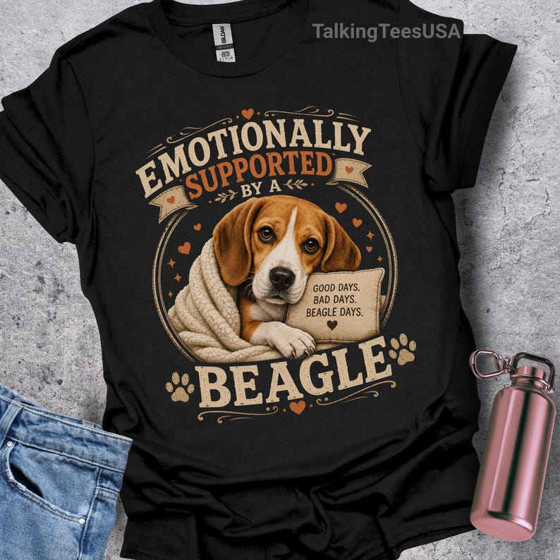 Może przedstawiać: Czarny t-shirt z grafiką przedstawiającą beagle'a owiniętego w beżowy koc. Napis głosi EMOTIONALLY SUPPORTED BY A BEAGLE oraz GOOD DAYS. BAD DAYS. BEAGLE DAYS. Wz&oacute;r zawiera małe pomarańczowe serduszka i odciski łap w okrągłej ramce.