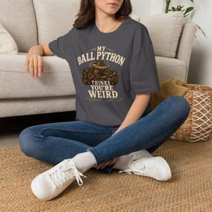 May include: A charcoal gray short-sleeve t-shirt featuring a graphic of a coiled ball python snake. The text printed in cream-colored lettering reads, "MY BALL PYTHON THINKS YOU'RE WEIRD," accompanied by small decorative flourishes and heart icons.