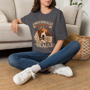 Może przedstawiać: Ciemnoszary t-shirt z grafiką przedstawiającą beagle'a owiniętego w koc. Napis głosi "EMOTIONALLY SUPPORTED BY A BEAGLE" oraz "GOOD DAYS, BAD DAYS, BEAGLE DAYS", a wok&oacute;ł ilustracji psa znajdują się małe serduszka i odciski łap.