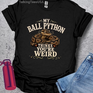 May include: A black short-sleeved t-shirt featuring a graphic of a coiled brown and black ball python. The text reads MY BALL PYTHON THINKS YOU'RE WEIRD in a beige font. The shirt includes a small brand tag at the inner neckline.