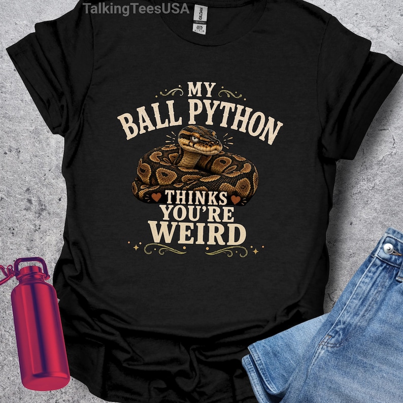 May include: A black short-sleeved t-shirt featuring a graphic of a coiled brown and black ball python. The text reads MY BALL PYTHON THINKS YOU'RE WEIRD in a beige font. The shirt includes a small brand tag at the inner neckline.