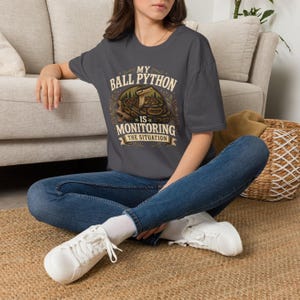 Puede incluir: Una camiseta gris carb&oacute;n de manga corta con un gr&aacute;fico de una pit&oacute;n bola enroscada en una rama y el texto "MY BALL PYTHON IS MONITORING THE SITUATION" en una fuente decorativa, combinada con vaqueros azules y zapatillas blancas.