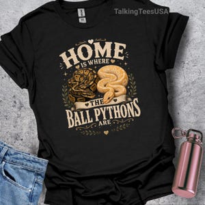 Peut inclure: Un t-shirt noir à manches courtes avec une impression graphique portant le texte "HOME IS WHERE THE BALL PYTHONS ARE". Le motif comprend des illustrations de deux pythons royaux, un brun foncé à motifs noirs et un beige clair, entourés de feuilles.