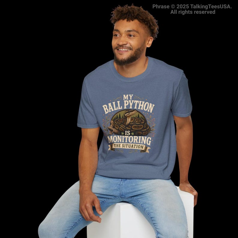 Puede incluir: Una camiseta de manga corta en color azul jaspeado con un gr&aacute;fico de una pit&oacute;n bola enroscada. El texto dice MY BALL PYTHON IS MONITORING THE SITUATION. El dise&ntilde;o incluye un marco circular con elementos forestales y una cinta inferior.