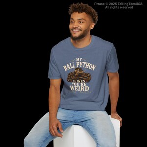 May include: A heather blue short-sleeve t-shirt featuring a graphic of a coiled brown ball python. The text printed in white and tan reads: MY BALL PYTHON THINKS YOU'RE WEIRD. The design is centered on the chest of the crew neck garment.