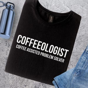 May include: Black crew neck t-shirt with the white text "COFFEEOLOGIST" and "COFFEE ASSISTED PROBLEM SOLVER". A blue water bottle and denim jeans are also visible.