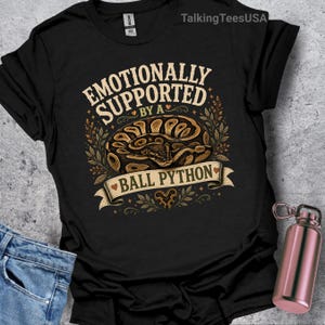 Peut inclure: Un t-shirt noir à manches courtes avec une illustration d'un python royal enroulé. Le texte "EMOTIONALLY SUPPORTED BY A BALL PYTHON" est écrit en lettres courbes. Le motif comprend des feuilles décoratives, des petits cœurs et une bannière.