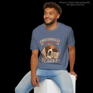 Może przedstawiać: Wrzosowoniebieski t-shirt z grafiką beagle'a owiniętego w koc. Tekst brzmi "EMOTIONALLY SUPPORTED BY A BEAGLE" oraz "GOOD DAYS, BAD DAYS, BEAGLE DAYS" z małą ikoną serca. Wz&oacute;r zawiera odciski łap i ozdobne linie wok&oacute;ł centralnego motywu.