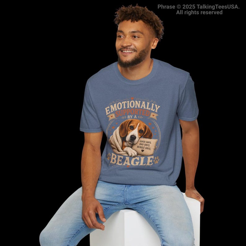 Może przedstawiać: Wrzosowoniebieski t-shirt z grafiką beagle'a owiniętego w koc. Tekst brzmi "EMOTIONALLY SUPPORTED BY A BEAGLE" oraz "GOOD DAYS, BAD DAYS, BEAGLE DAYS" z małą ikoną serca. Wz&oacute;r zawiera odciski łap i ozdobne linie wok&oacute;ł centralnego motywu.