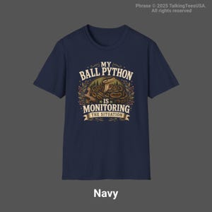 Puede incluir: Una camiseta de manga corta azul marino con un gr&aacute;fico de una pit&oacute;n bola enroscada en una rama. El texto dice MY BALL PYTHON IS MONITORING THE SITUATION, rodeado de follaje decorativo y un dise&ntilde;o de pancarta en tonos tierra.