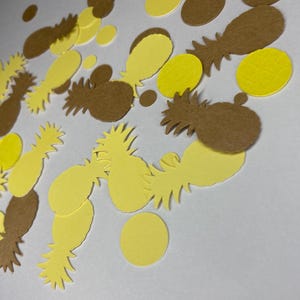 May include: Overhead shot of yellow and brown confetti in the shape of pineapples and circles. The confetti is scattered across a white surface, creating a playful and festive look. The pineapple shapes have detailed cutouts.