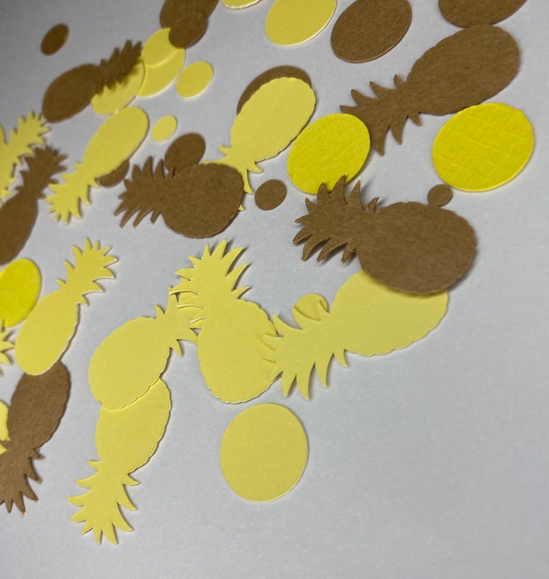 May include: Overhead shot of yellow and brown confetti in the shape of pineapples and circles. The confetti is scattered across a white surface, creating a playful and festive look. The pineapple shapes have detailed cutouts.