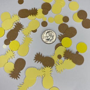 May include: A collection of yellow and brown pineapple and circle-shaped confetti scattered on a white surface. A U.S. dime is in the center of the confetti. The confetti is made of paper and is likely used for party decorations.