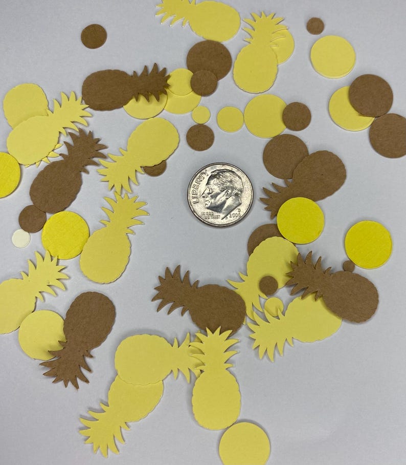 May include: A collection of yellow and brown pineapple and circle-shaped confetti scattered on a white surface. A U.S. dime is in the center of the confetti. The confetti is made of paper and is likely used for party decorations.