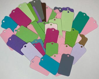 May include: A collection of colorful gift tags in various shades of pink, green, purple, and gray. Each tag has a rounded top with a small hole for stringing. The tags are arranged in a heart-like shape against a white background.