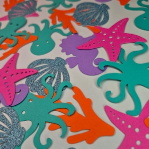 May include: A selection of colourful, die-cut ocean-themed shapes. The assortment includes teal octopuses, coral, starfish, seashells, and seahorses in shades of pink, orange, purple, and teal. Some shapes have glitter accents.