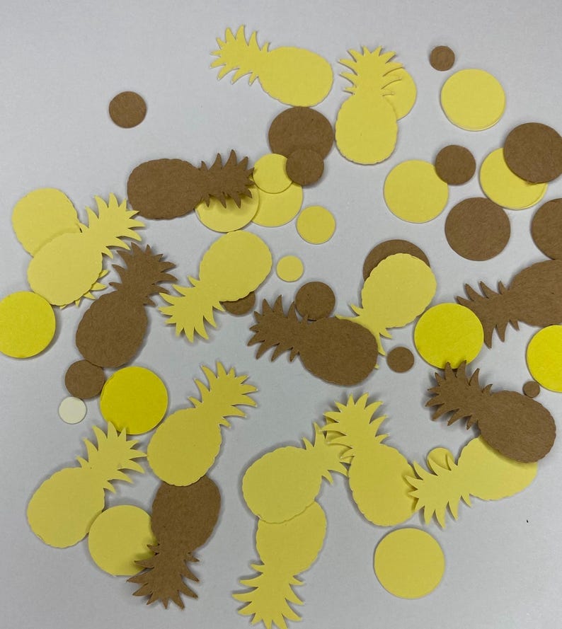 May include: Overhead shot of scattered yellow and brown paper cutouts. The cutouts include pineapple shapes and circular shapes. The pineapples are in various shades of yellow and brown. The circles are yellow and brown.