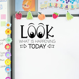 May include: A white dry-erase board with the text "LOOK WHAT IS HAPPENING TODAY" in black with large cartoon eyes. The board is decorated with colorful flowers and other stickers.