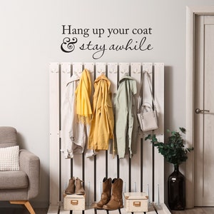 May include: White wooden coat rack with hooks and a sign that reads "Hang up your coat & stay awhile". There are four coats hanging on the rack, including a yellow jacket, a white jacket, a green jacket, and a scarf. There are also two pairs of brown boots and a brown purse on the rack.