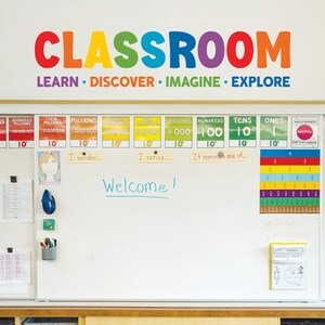 May include: A white board with the word "CLASSROOM" in rainbow letters and the words "LEARN DISCOVER IMAGINE EXPLORE" underneath. The board is decorated with colorful charts and posters, including a place value chart, a fraction chart, and a welcome sign.